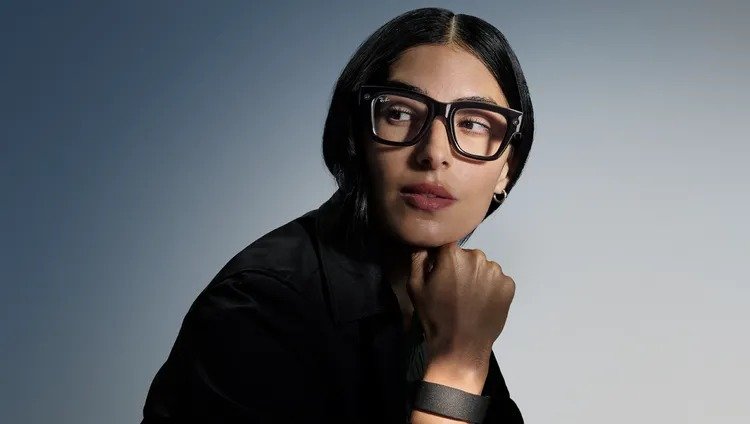 Woman with dark hair wearing large black Ray-Ban glasses, looking thoughtful.