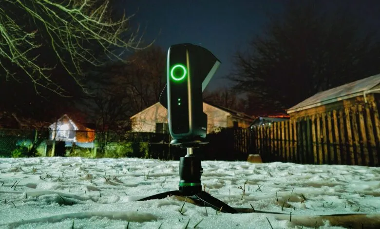 Black 3D scanner on tripod in snowy yard at night with green light.