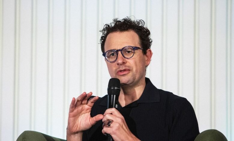 Man with glasses speaking into a microphone, gesturing with his hand.