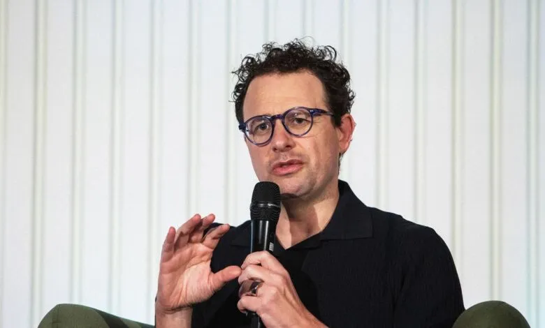 Man with glasses speaking into a microphone, gesturing with his hand.