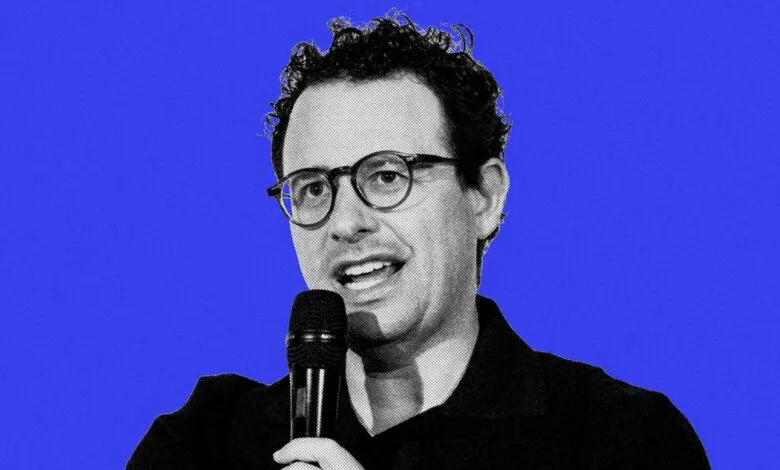 Nathan Fielder speaking into a microphone against a blue background.