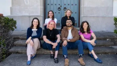 Six smiling people sit on stone steps in front of a building with a blue door.