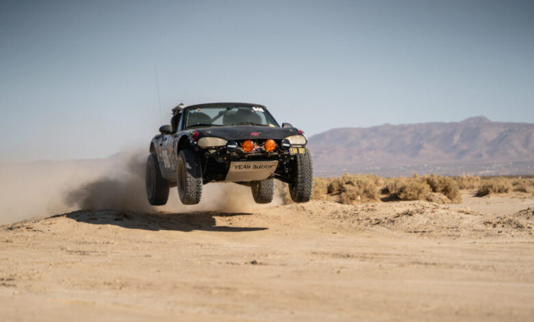 Off-road Miata jumps over a sand dune, dust billowing behind it.