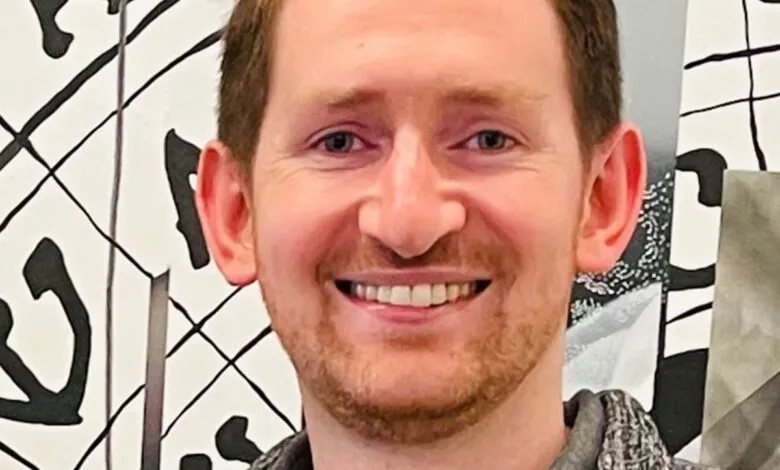 Smiling man with reddish hair in front of a black and white patterned background.