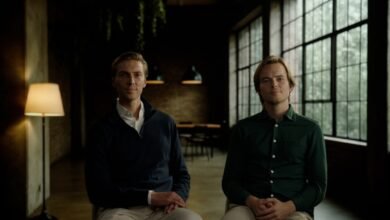 Two men sit side-by-side in a dimly lit room with large windows.