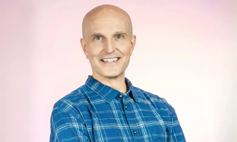 Portrait of a smiling bald man wearing a blue plaid shirt against a pink background.