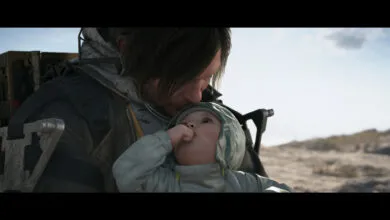 Man with dark hair holds a baby wearing a hooded jacket in an outdoor setting.