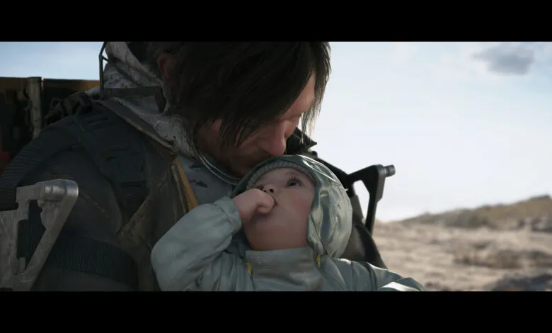 Man with dark hair holds a baby wearing a hooded jacket in an outdoor setting.