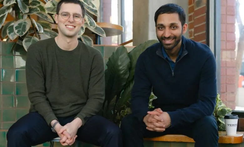 Two men sit side-by-side smiling in a bright, plant-filled space.