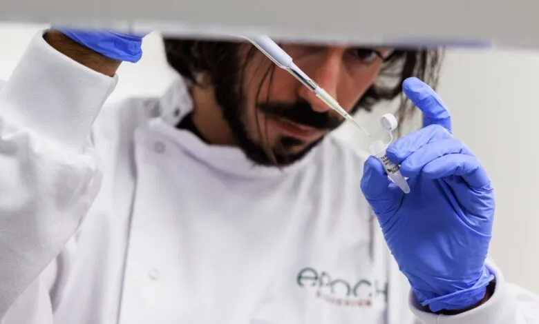 Scientist in lab coat using pipette to transfer liquid into vial.