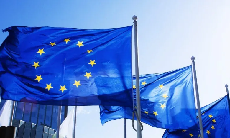 Several EU flags waving against a bright blue sky and modern building.