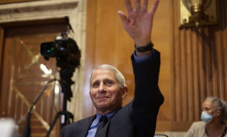 Dr. Anthony Fauci raises hand during a hearing, wearing a suit.