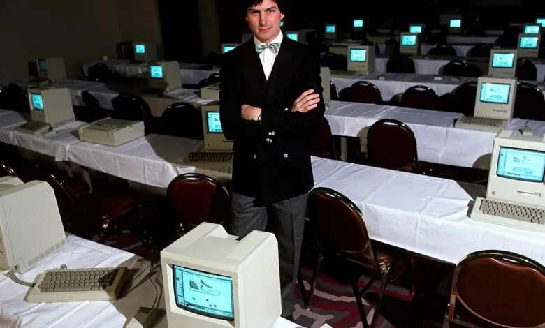 Steve Jobs stands in a room full of vintage Macintosh computers.
