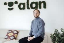 Man with glasses sits on a couch in front of a moss-covered 'alan' sign.