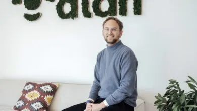 Man with glasses sits on a couch in front of a moss-covered 'alan' sign.