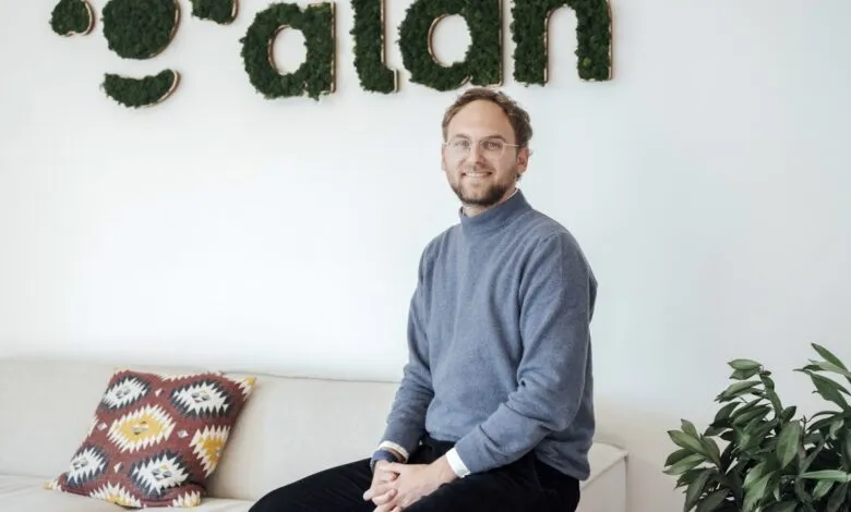 Man with glasses sits on a couch in front of a moss-covered 'alan' sign.