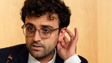 Man with glasses adjusts ear during a meeting, looking thoughtful.