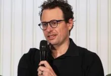 Nathan Fielder speaking into a microphone with a white background.