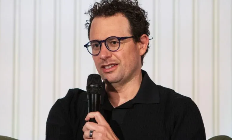 Nathan Fielder speaking into a microphone with a white background.