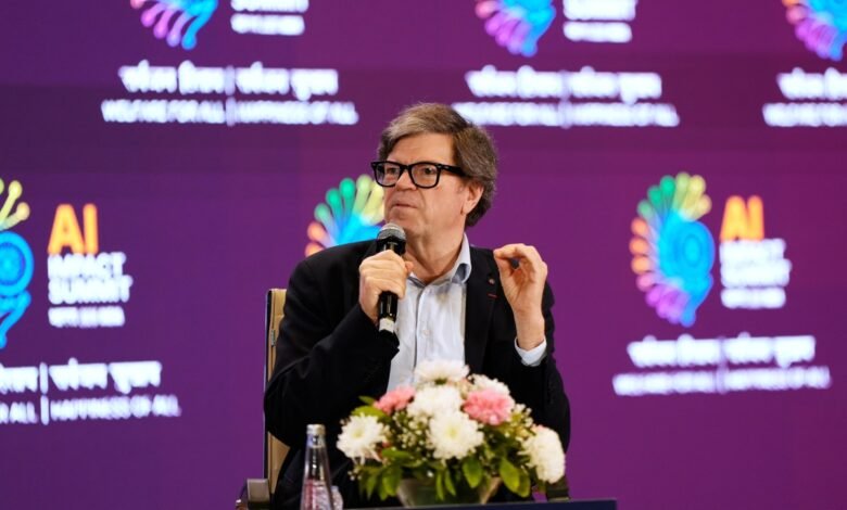 Man with glasses speaks at AI Impact Summit, holding microphone.