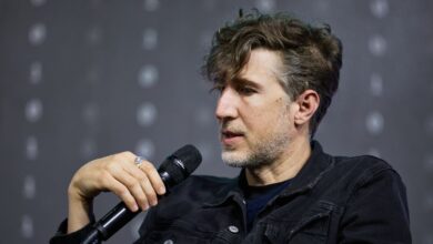 Man with curly hair holds microphone, wearing denim jacket.