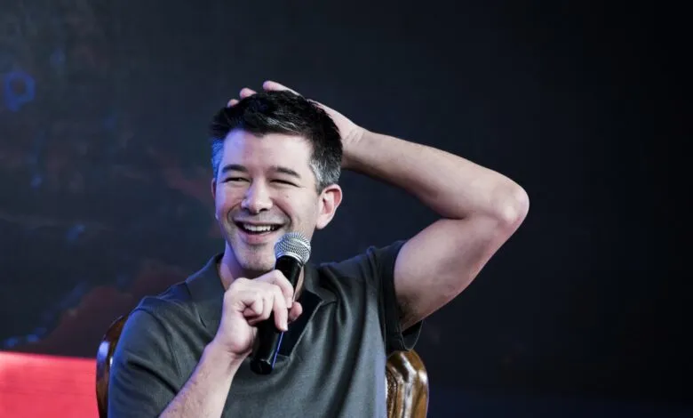 Travis Kalanick smiles, holding microphone, hand on head.