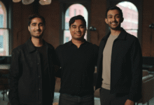 Three young men smiling indoors, posing in front of brick walls and windows.