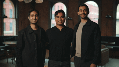 Three young men smiling indoors, posing in front of brick walls and windows.