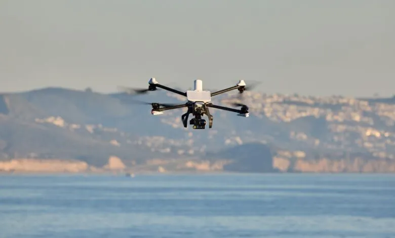 White and black drone hovers over blue water with blurred coastline in background.