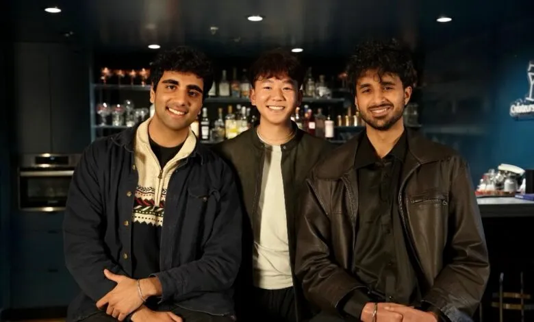 Three young men smiling in front of a bar with bottles and glasses.
