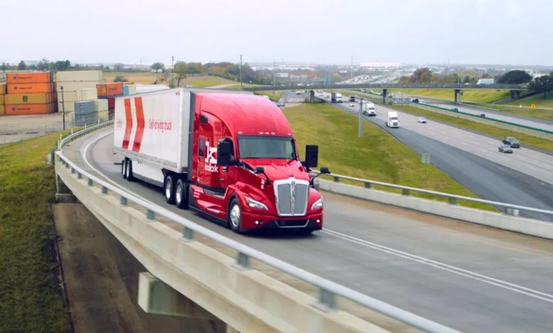 Red Kodiak self-driving truck on highway overpass with shipping containers in background.