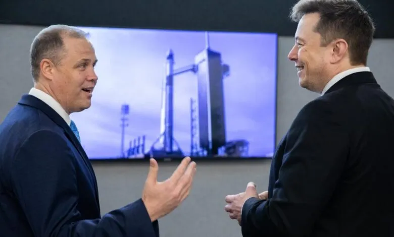 Two men in suits, including Elon Musk, converse in front of a rocket display.