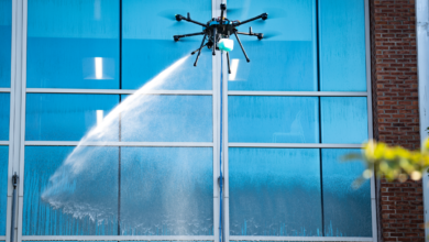 Drone cleaning tall building's blue glass windows with a water spray.