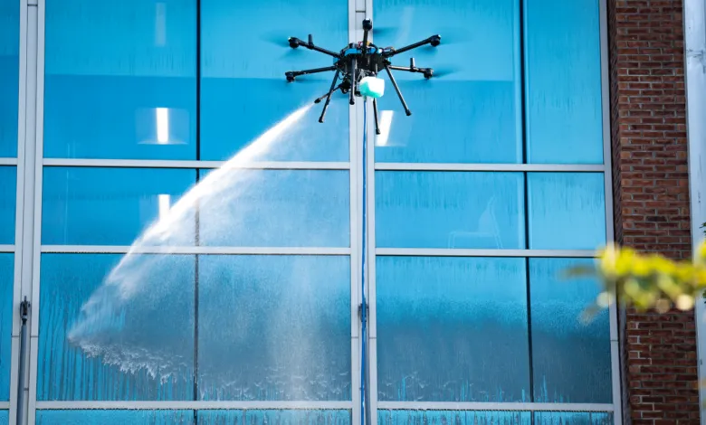 Drone cleaning tall building's blue glass windows with a water spray.
