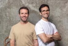 Two smiling men stand side-by-side against a textured gray wall.
