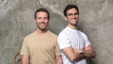 Two smiling men stand side-by-side against a textured gray wall.