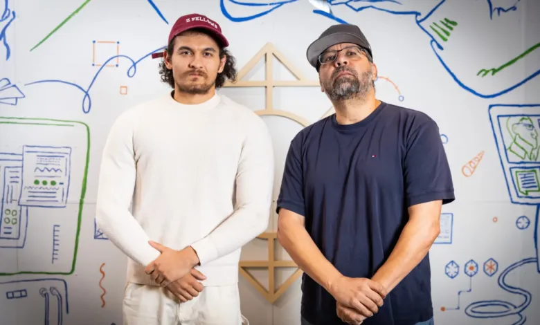 Two men stand in front of a wall with drawings, one wearing a 'Z Fellows' cap.