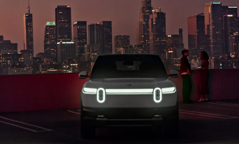 Rivian R2 electric SUV with illuminated headlights against a city skyline at dusk.