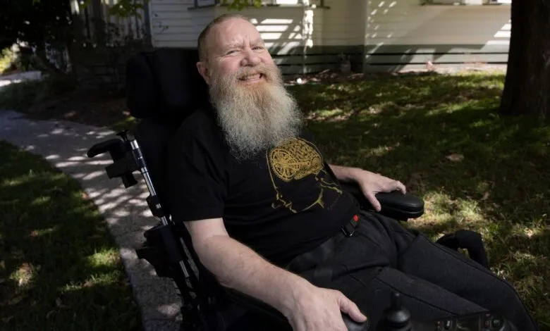 Man with beard in wheelchair smiles outdoors on sunny day.
