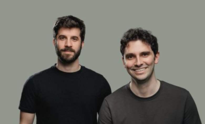 Two men stand side-by-side against a gray background, smiling.