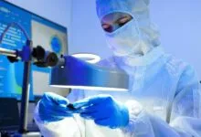 Technician in clean suit inspecting semiconductor wafer under bright light.