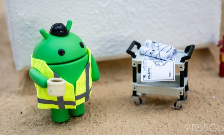 Android figure in construction vest holding coffee next to blueprints.