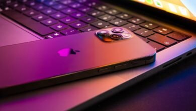 iPhone resting on a MacBook keyboard, illuminated with purple and orange light.