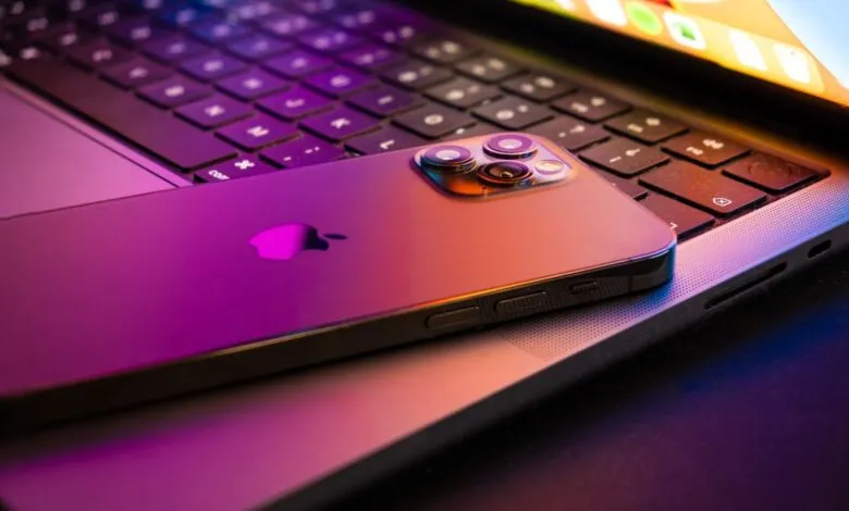 iPhone resting on a MacBook keyboard, illuminated with purple and orange light.