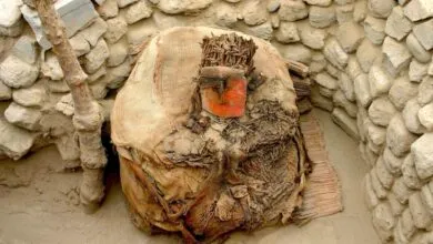Ancient burial bundle with a red mask, surrounded by stone walls and sand.