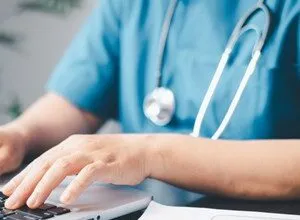 Healthcare worker in scrubs types on laptop with stethoscope around neck.