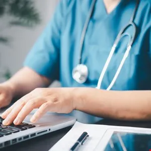 Healthcare worker in scrubs types on laptop with stethoscope around neck.