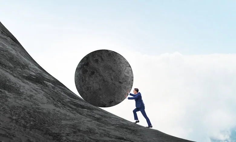 Man in suit struggles to push a large stone uphill against a cloudy sky.