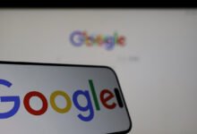 Smartphone displays Google logo in front of a blurred Google webpage on a monitor.