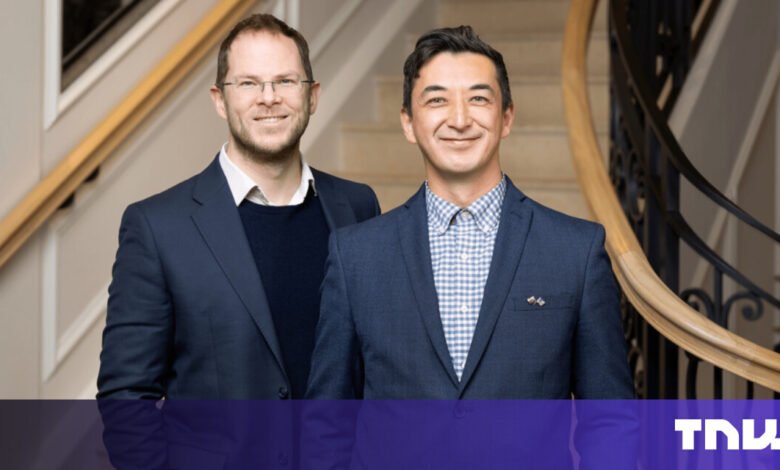 Two men in suits stand smiling on a staircase.